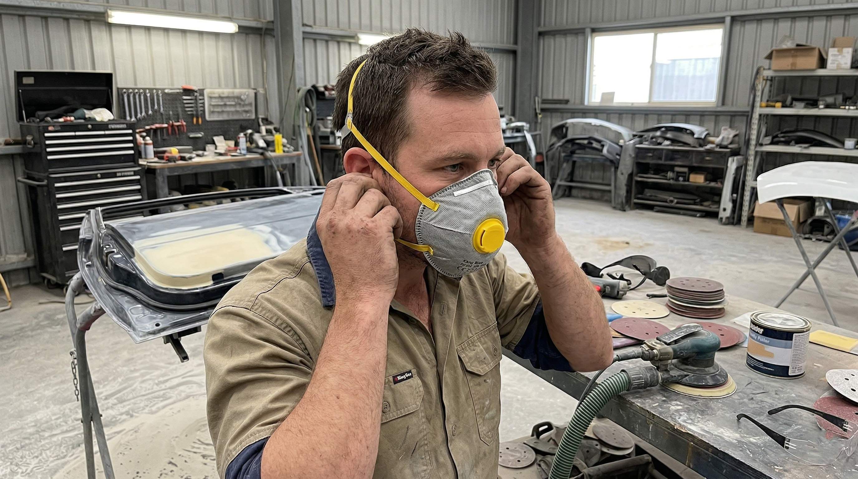 Tradesperson fitting a P2 dust mask before sanding a vehicle panel in a workshop.
